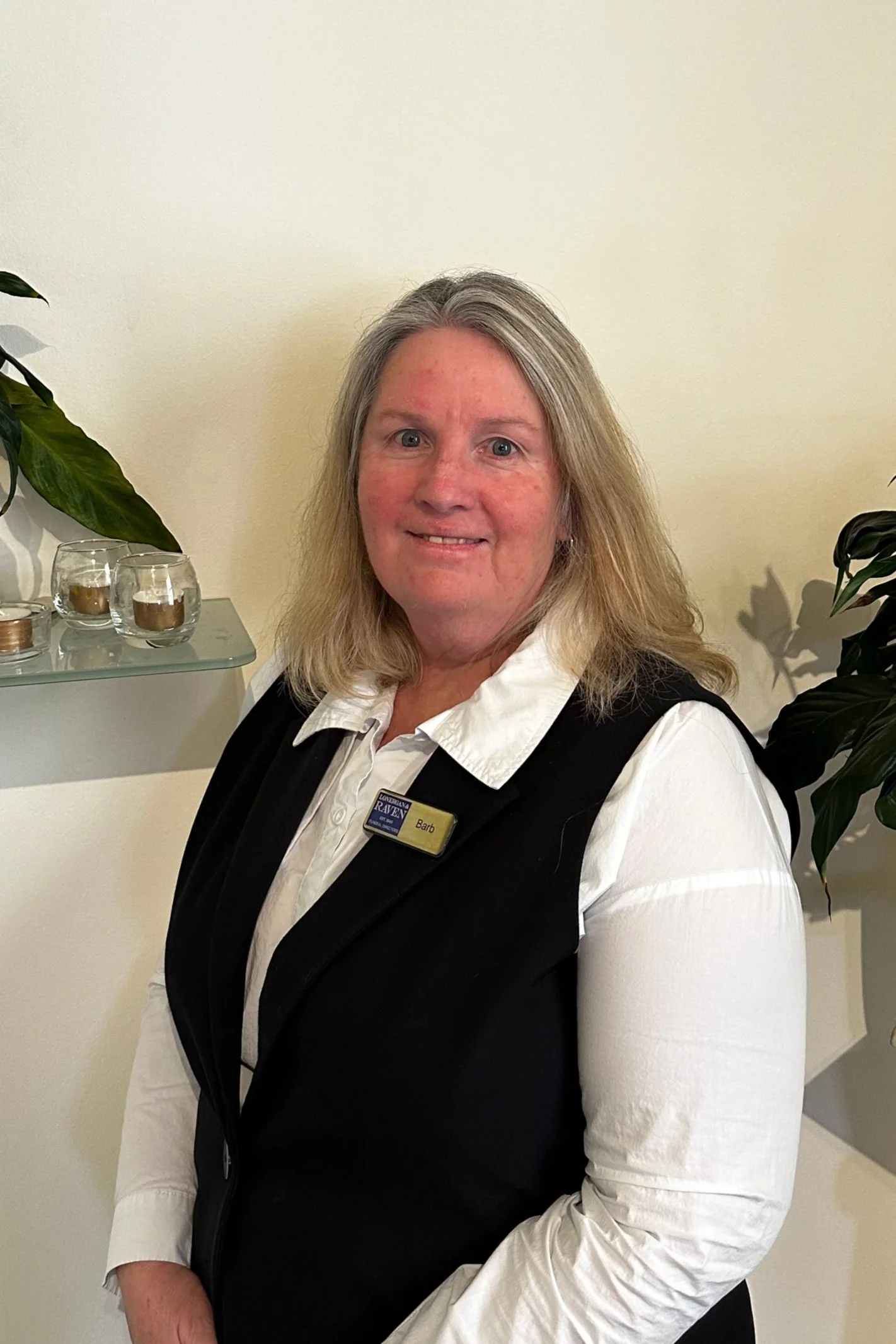 A woman with blonde hair in a uniform with a name tag that reads 'Barb' standing indoors next to houseplants and glass candle holders on a wall shelf.