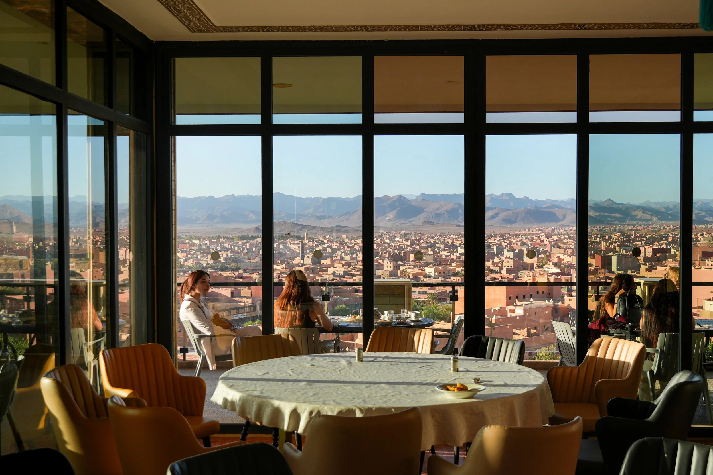 A restaurant interior with a large round table covered with a white tablecloth, surrounded by beige and black chairs. Large glass windows provide a view of a cityscape and mountains in the distance. Several women are sitting at tables near the window