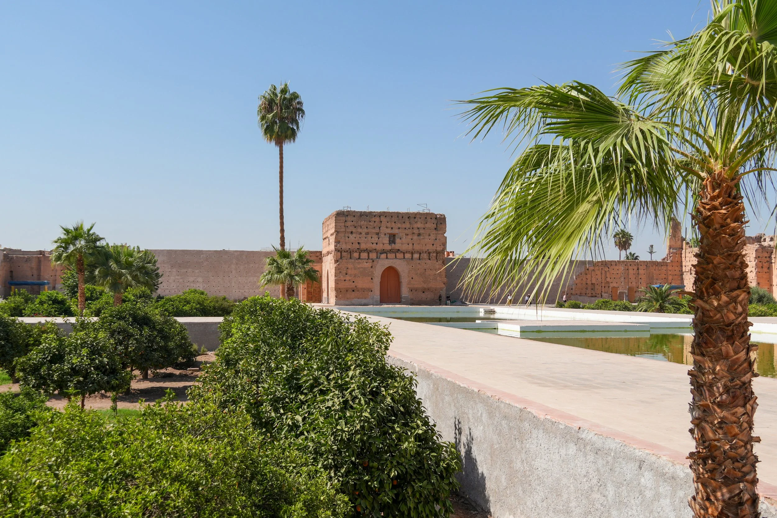 A historical brick fort wall with an arched wooden door, surrounded by palm trees, bushes, and a pool of water, under a clear blue sky.