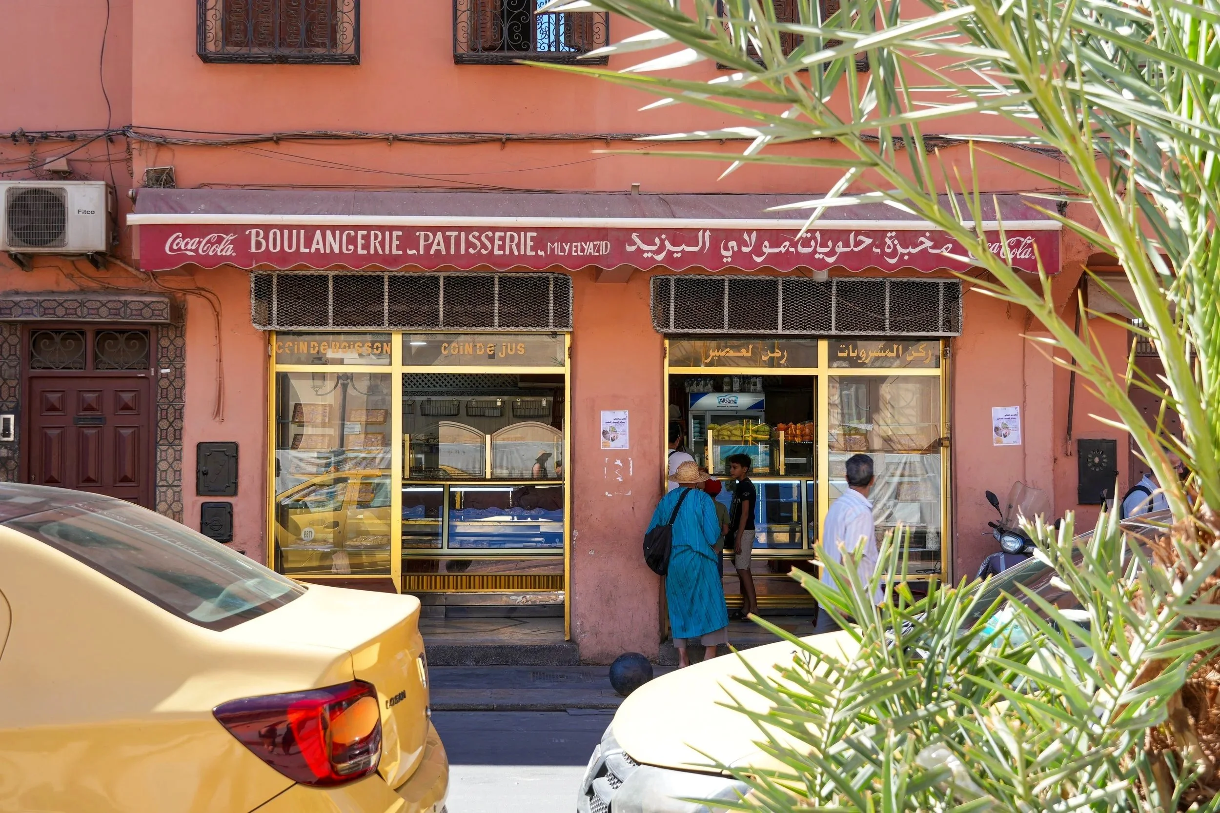 A bakery with a maroon sign displaying 'BOULANGERIE - PATISSERIE' in French and Arabic, with yellow-framed windows showing baked goods inside. Several people are standing outside, and a yellow car is parked in front. Green foliage partially obscures 