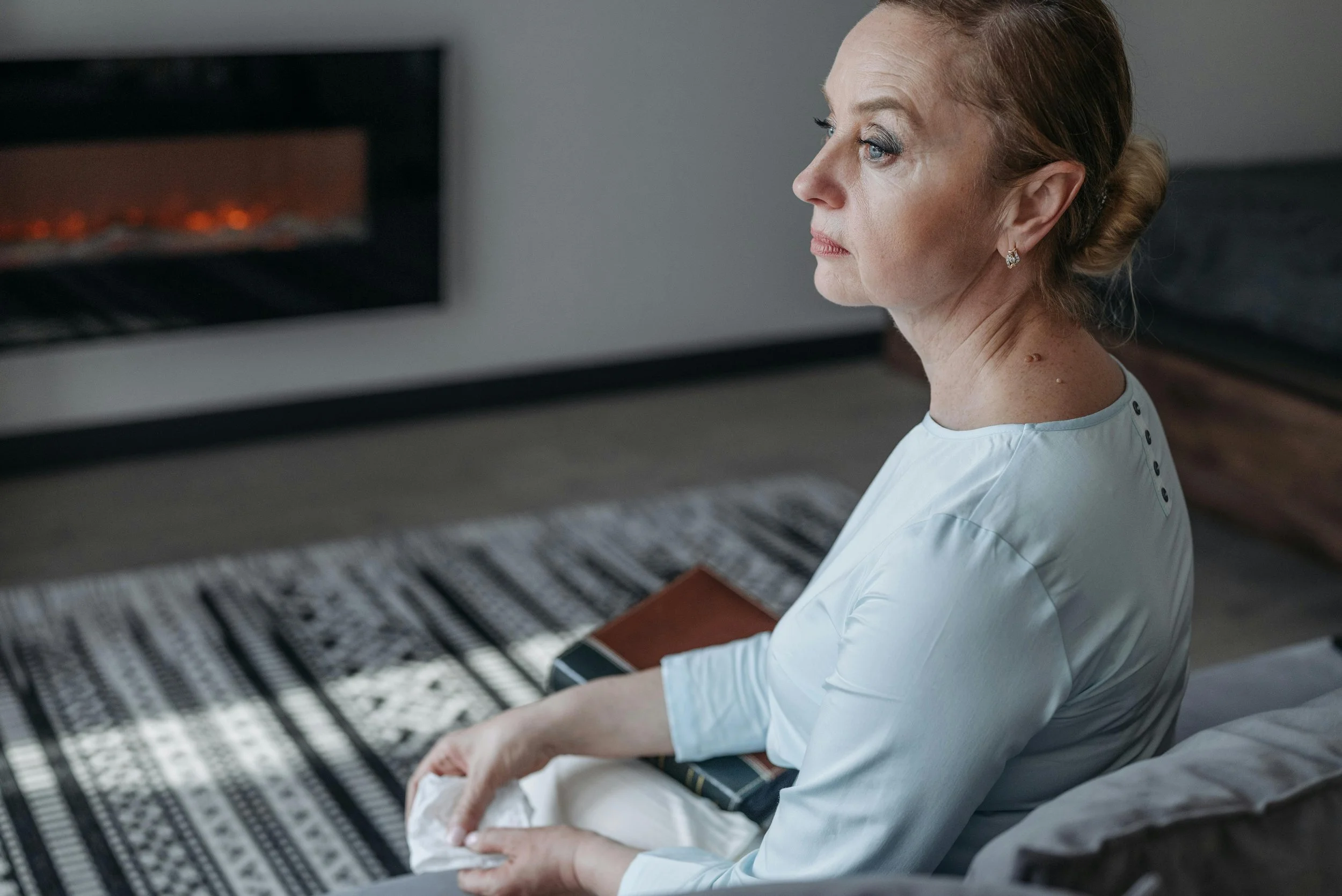 a woman sits on a couch in front of a fire with a book on her lap, looking pensively into the distance