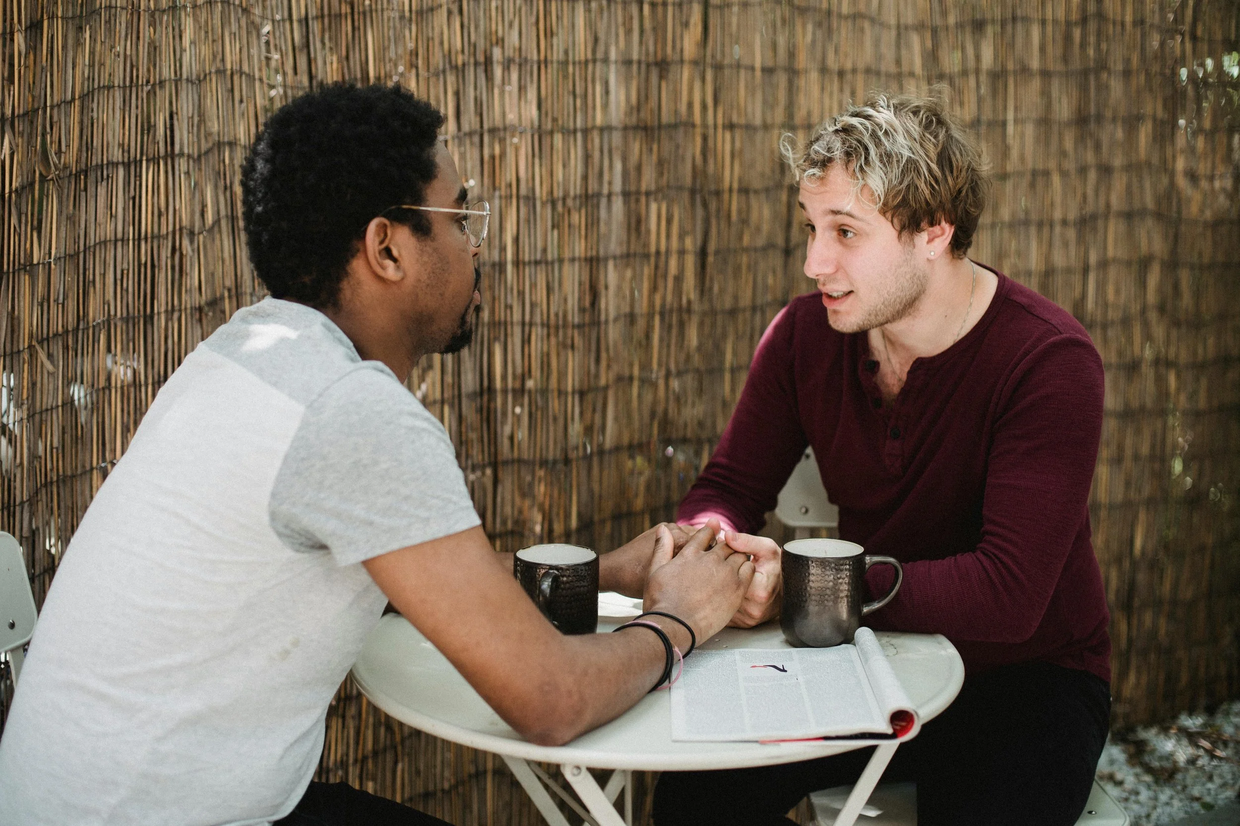 two men are sitting across from each other at a small table with mugs and an open magazine on it, they areholding hands