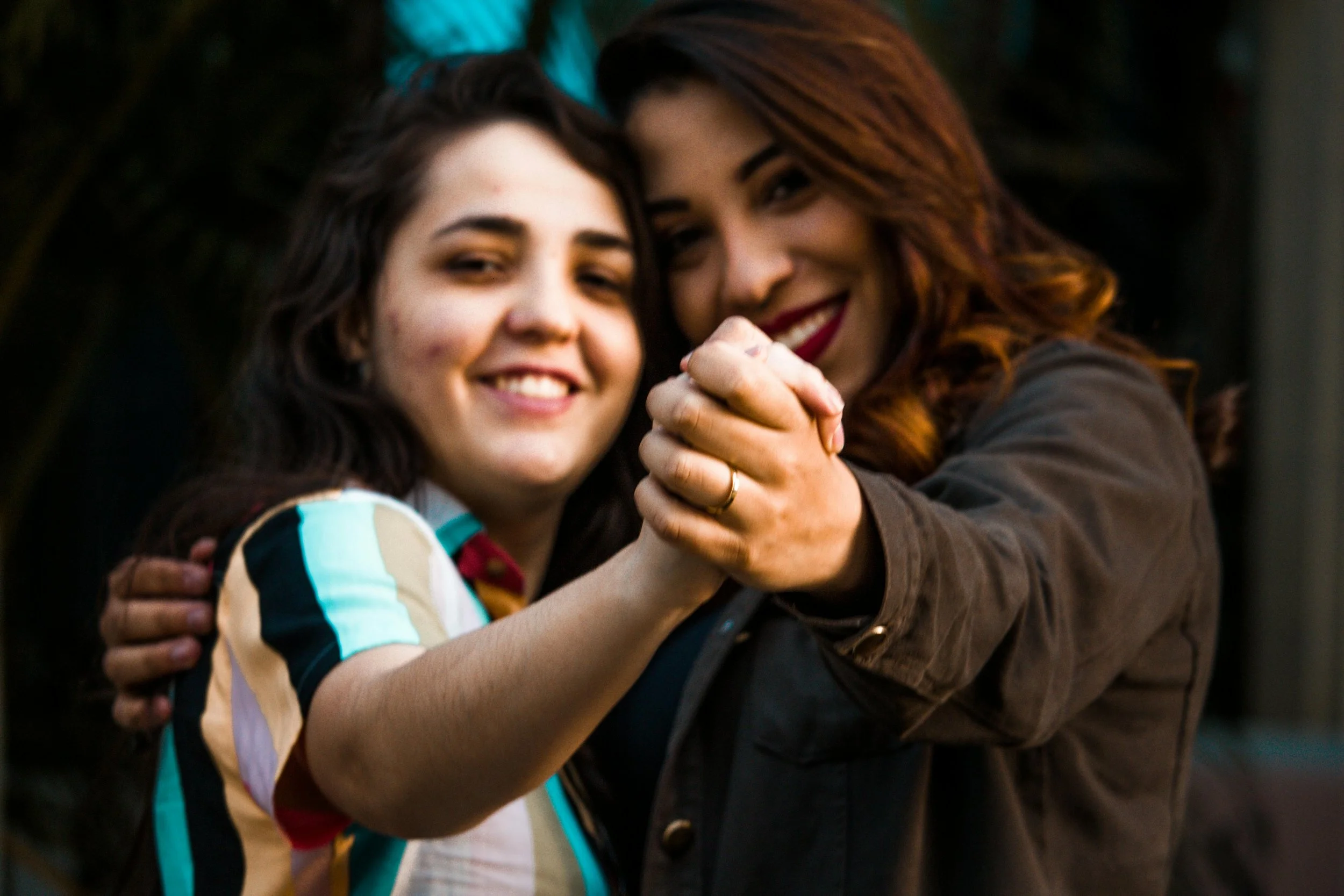 two women have their arms around each other and are holding hands, smiling towards the camera.