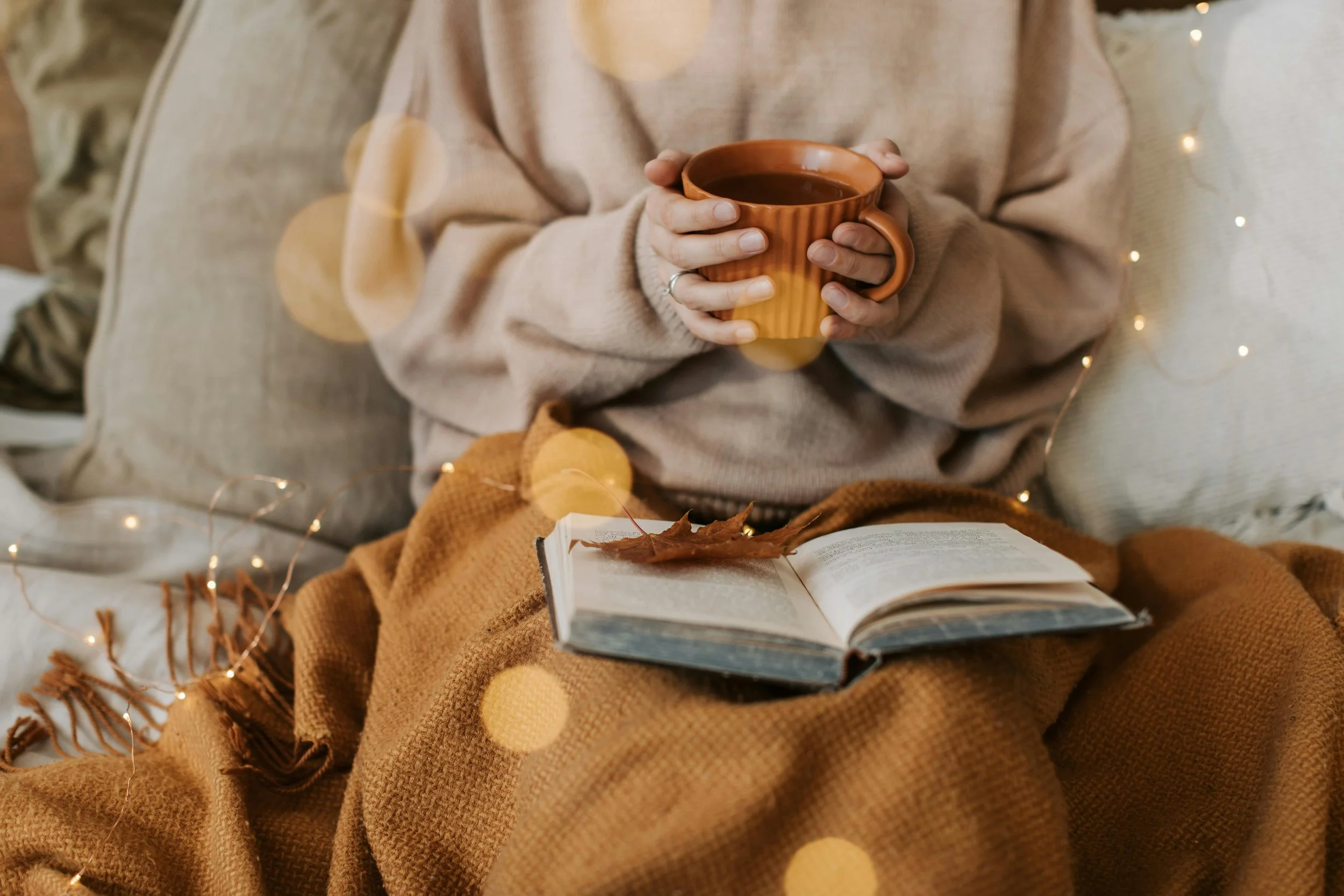 a person is sitting reading a book with twinkle lights across their lap and a mug in their hands.