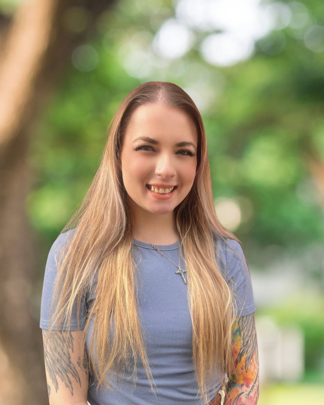 a young woman with long blond hair, a blue shirt and tattoos on her arms stands against a brick wall smiling