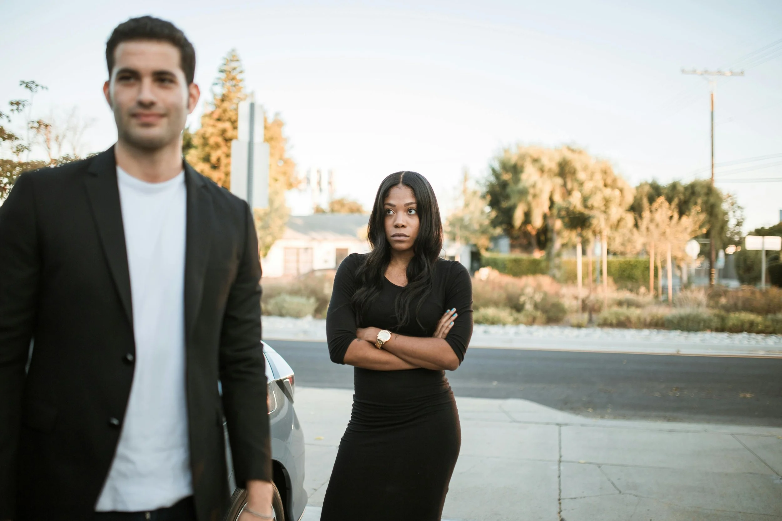 woman wearing a long black dress stands with her arms crossed and a displeased expression on her face while a man in a black blazer is walking in front of her smiling slightly