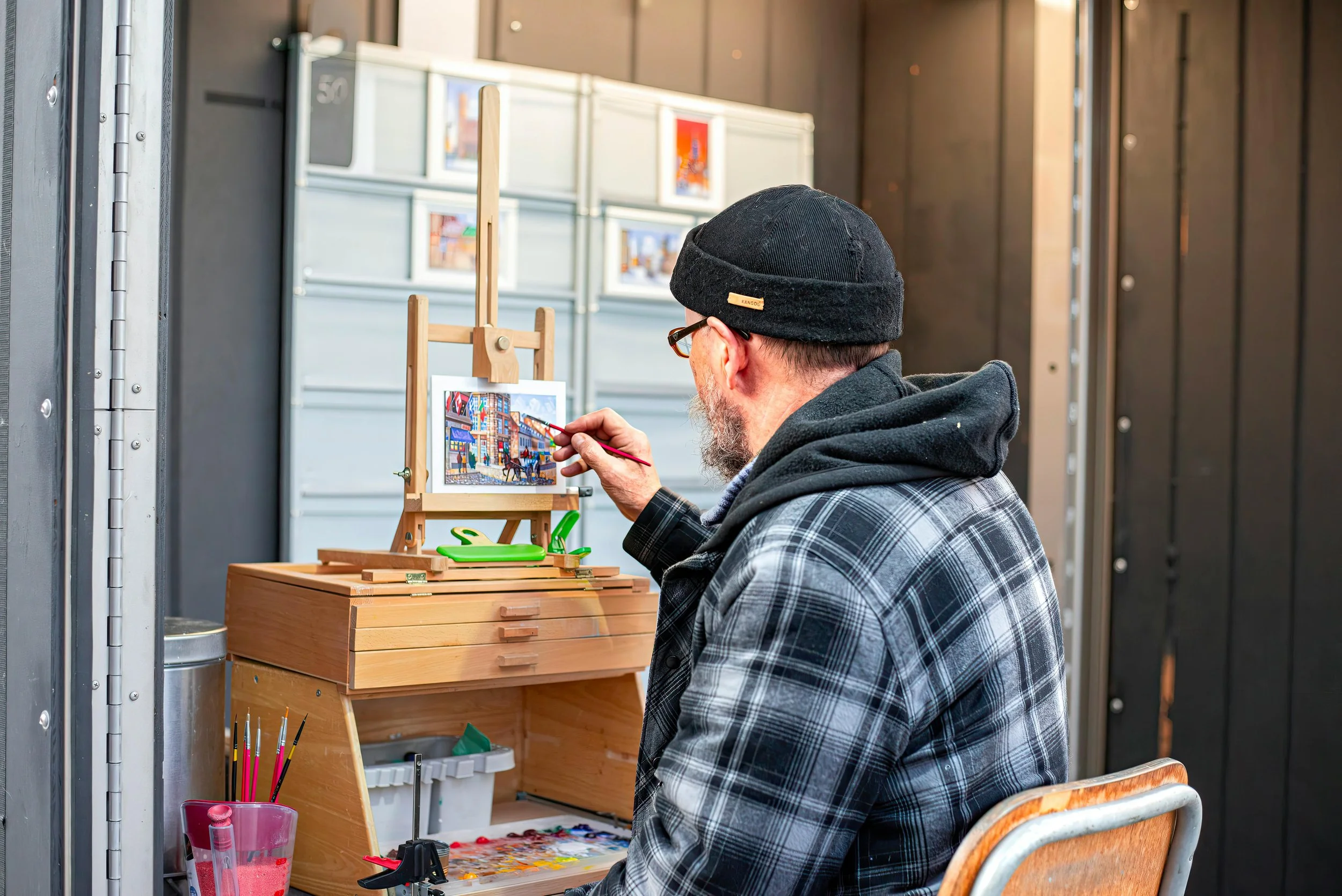 a man sits in front of a small easel with paints and paintbrushes in front of him, painting a small city scene