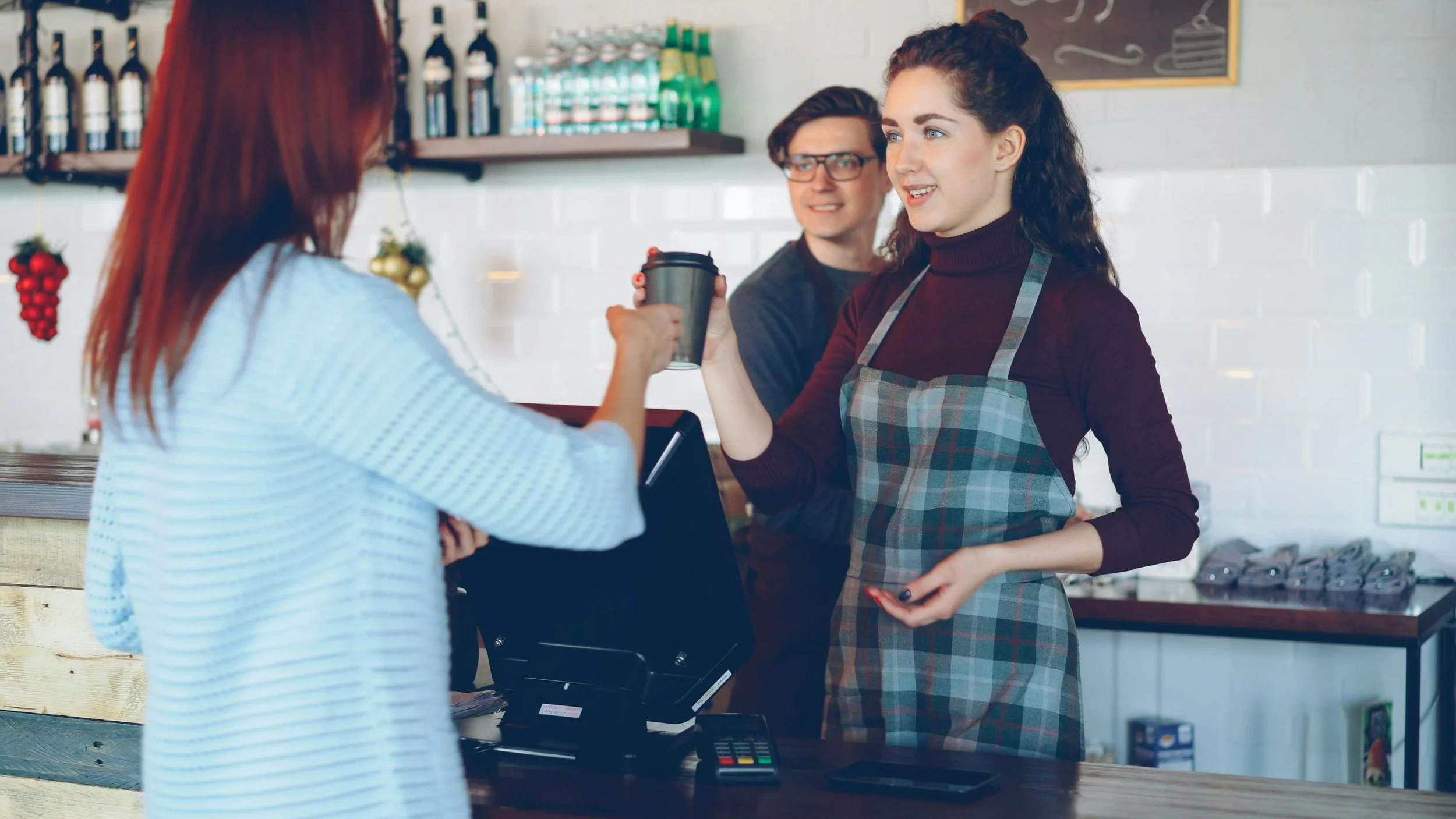 two women at either side of a barista counter are smiling each other, the cashier is handing the other woman a coffee and there is another barista in the background