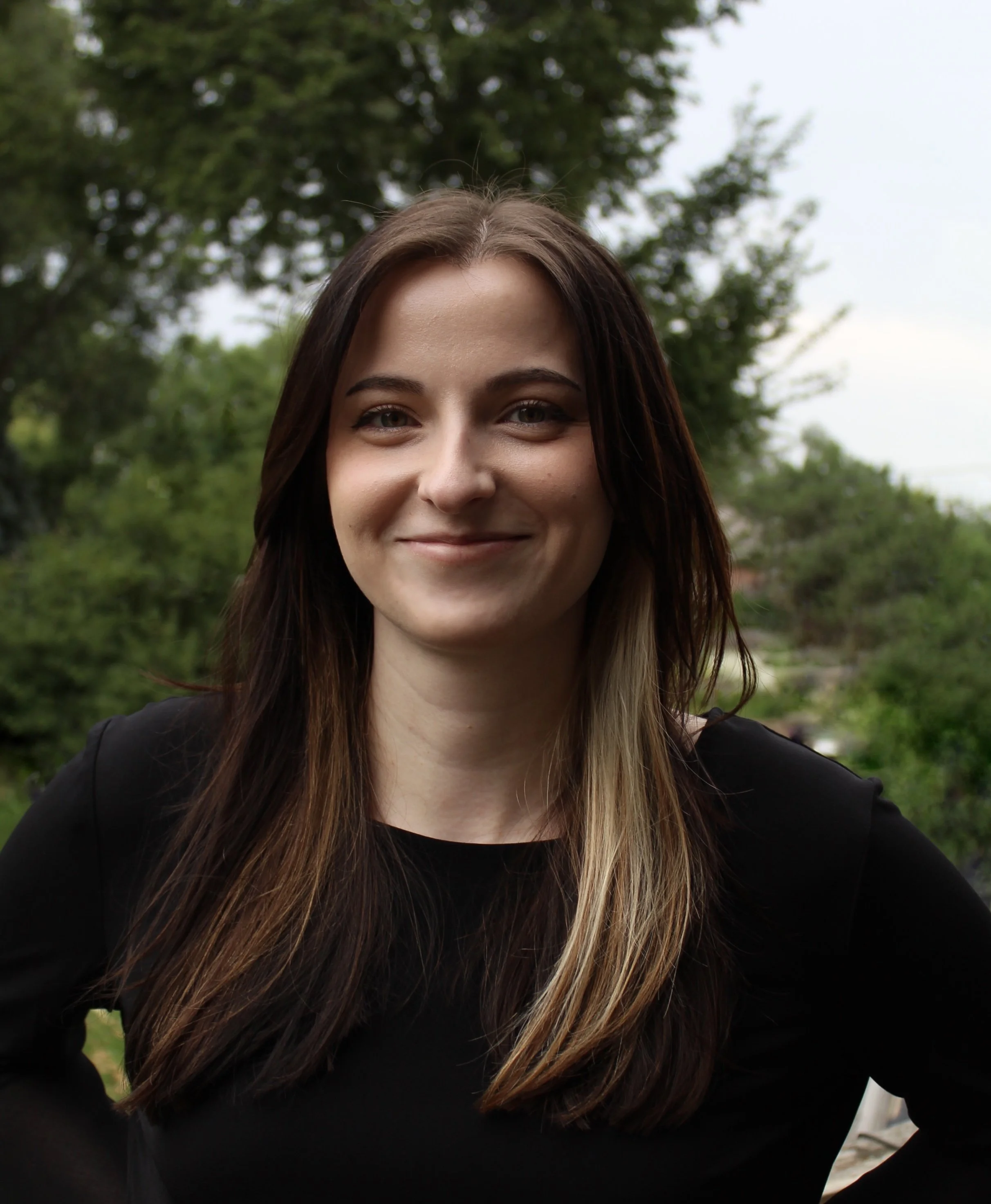 headshot of young woman with long brown hair and black shirt on standing against a wooded background, smiling