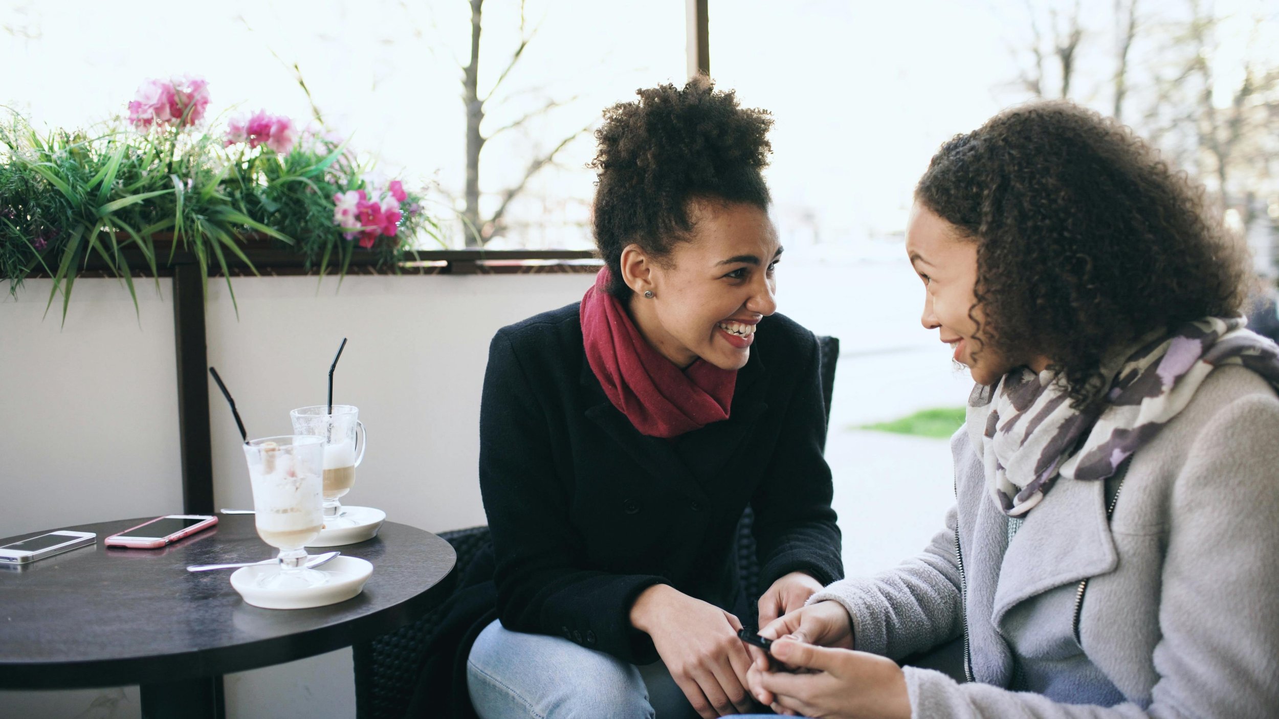 two women are looking into each others eyes talking and smiling outside on a patio with their phone and two empty milkshakes on the table next to them