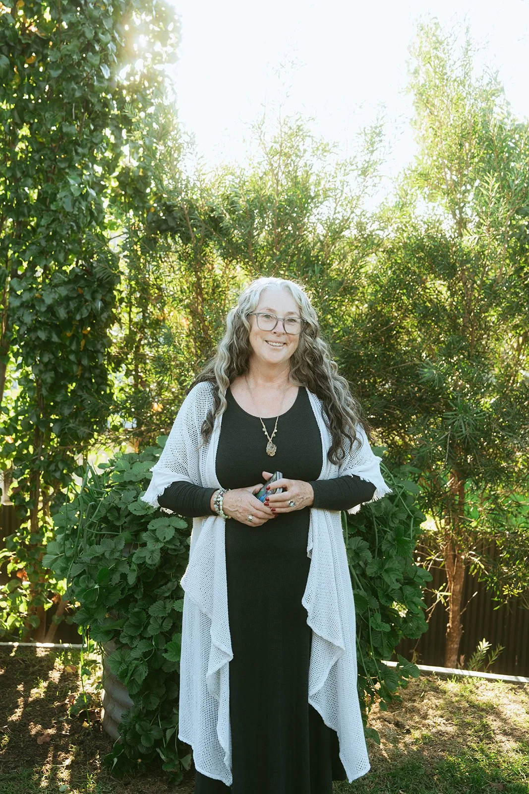 A smiling woman with dark hair, glasses, and wearing a floral dress, sitting at a table outdoors near a metal wall or trailer on a sunny day.