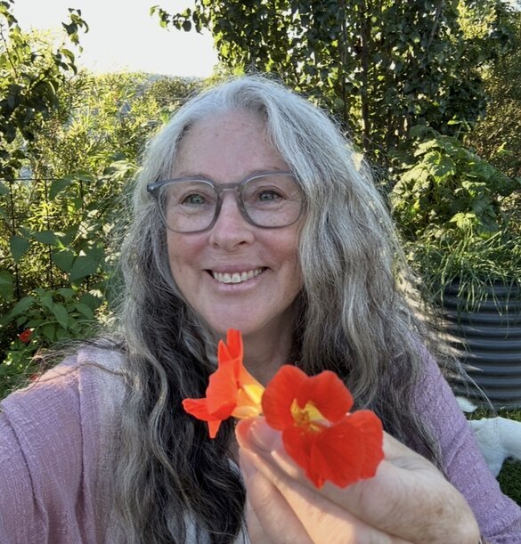 A smiling woman with dark hair, glasses, and wearing a floral dress, sitting at a table outdoors near a metal wall or trailer on a sunny day.