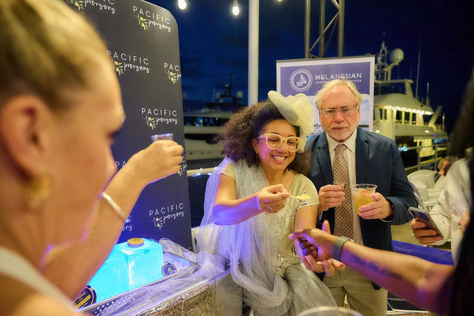 People socializing at an event at night, with a woman smiling and wearing a large yellow hat and glasses, and a man in a suit holding a drink.
