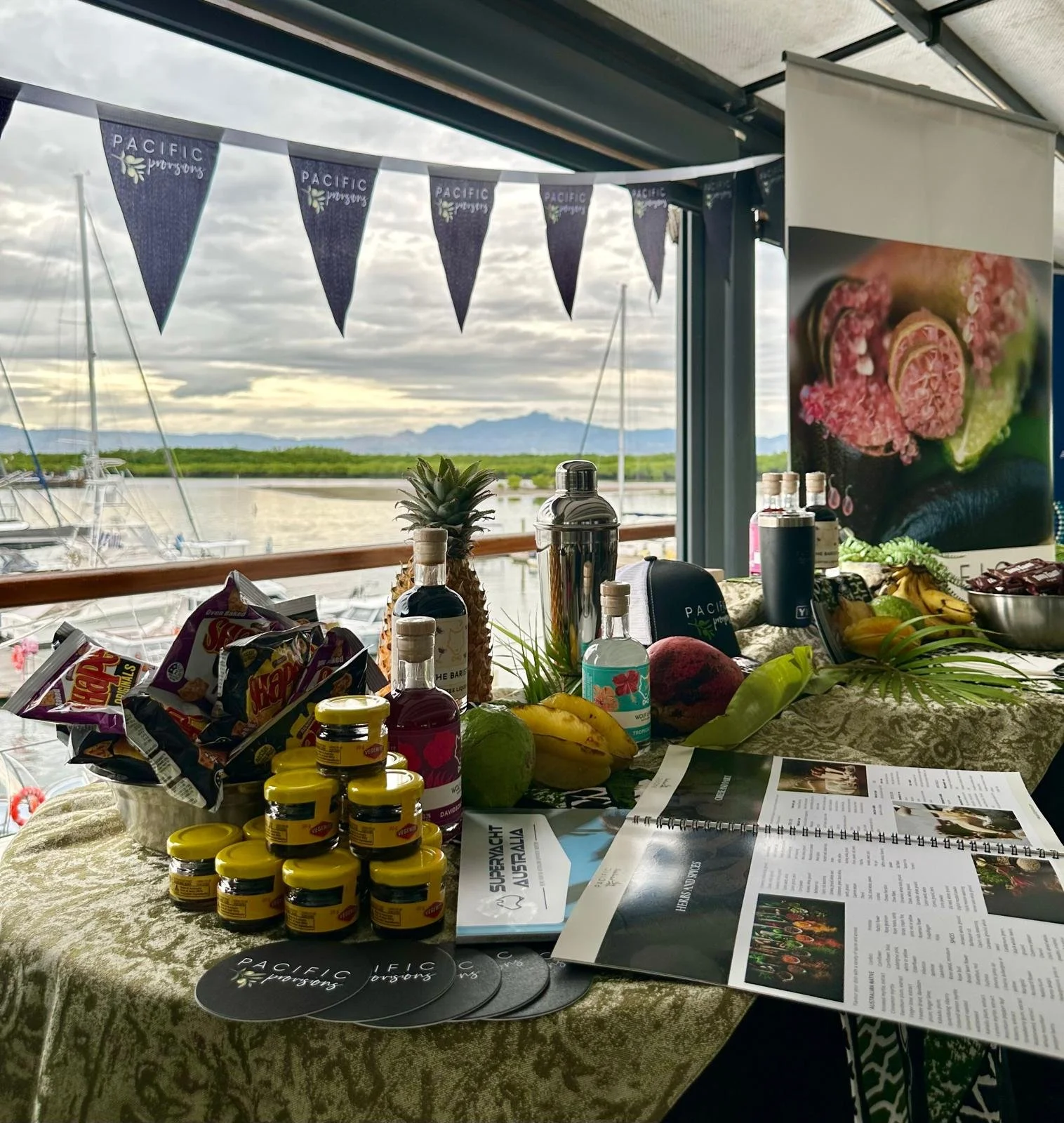 A table covered with snacks, bottles, and pamphlets, overlooking a marina with boats and a cloudy sky in the background. There is a banner with 'Pacific Provisions"' and a poster displaying food items.