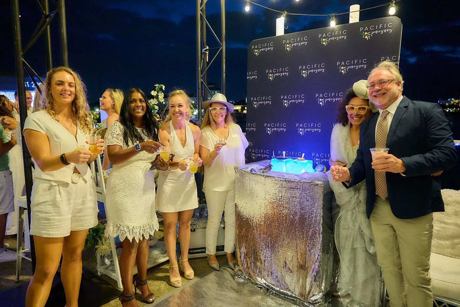 Group of six people celebrating at an evening event, standing around a shimmering bar with a backdrop featuring the words "Pacific Prorings" repeatedly. They are holding drinks and smiling, with some wearing light-colored or white clothing, and standing under string lights.