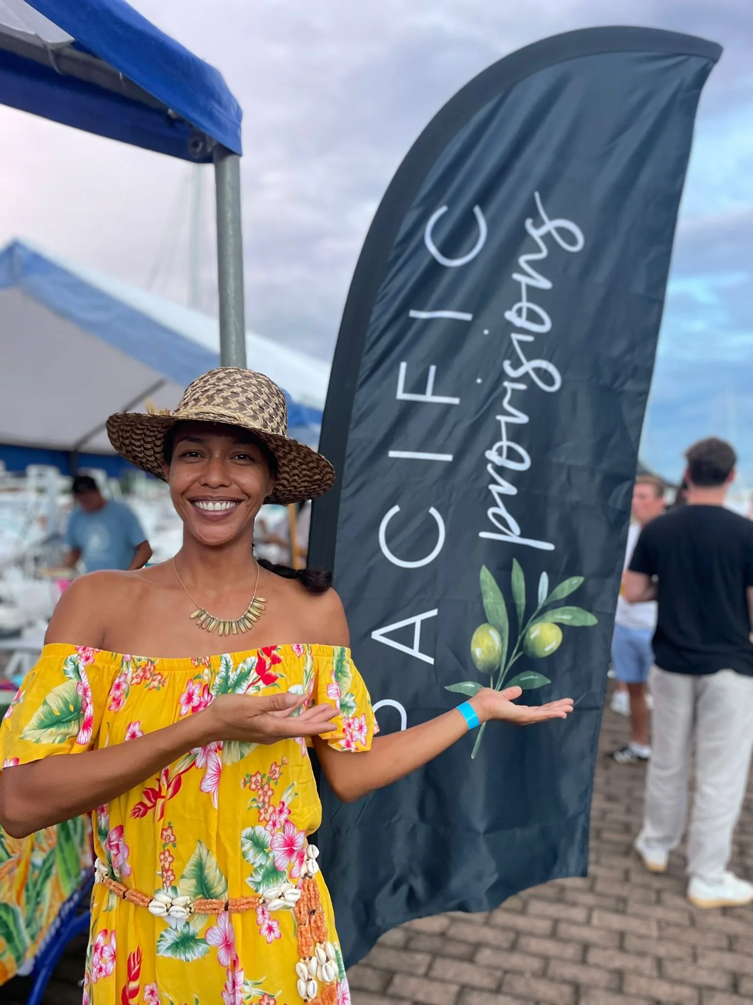 A smiling woman wearing a yellow tropical dress with a floral pattern and shell belt, holding a sign with an olive branch illustration and the text 'CULTIVATING CALIFORNIA.' She has on a straw hat and is at an outdoor event, with a blue sky and people in the background.