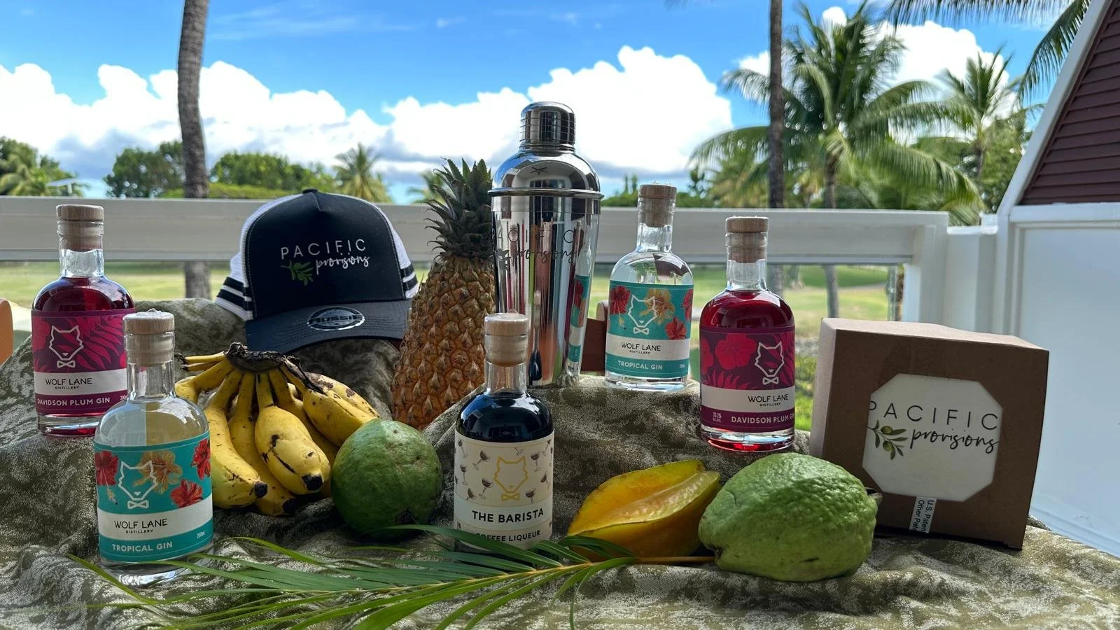 Assortment of spirits, tropical fruits including bananas, pineapple, and guava, a Pacific Provisions hat, a stainless steel cocktail shaker, and a box of Pacific Provisions products on a table outdoors with palm trees and a blue sky in the background.