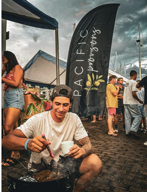 Young man cooking on a portable grill at an outdoor event with a Pacific IC banner in the background, and people standing and talking around.