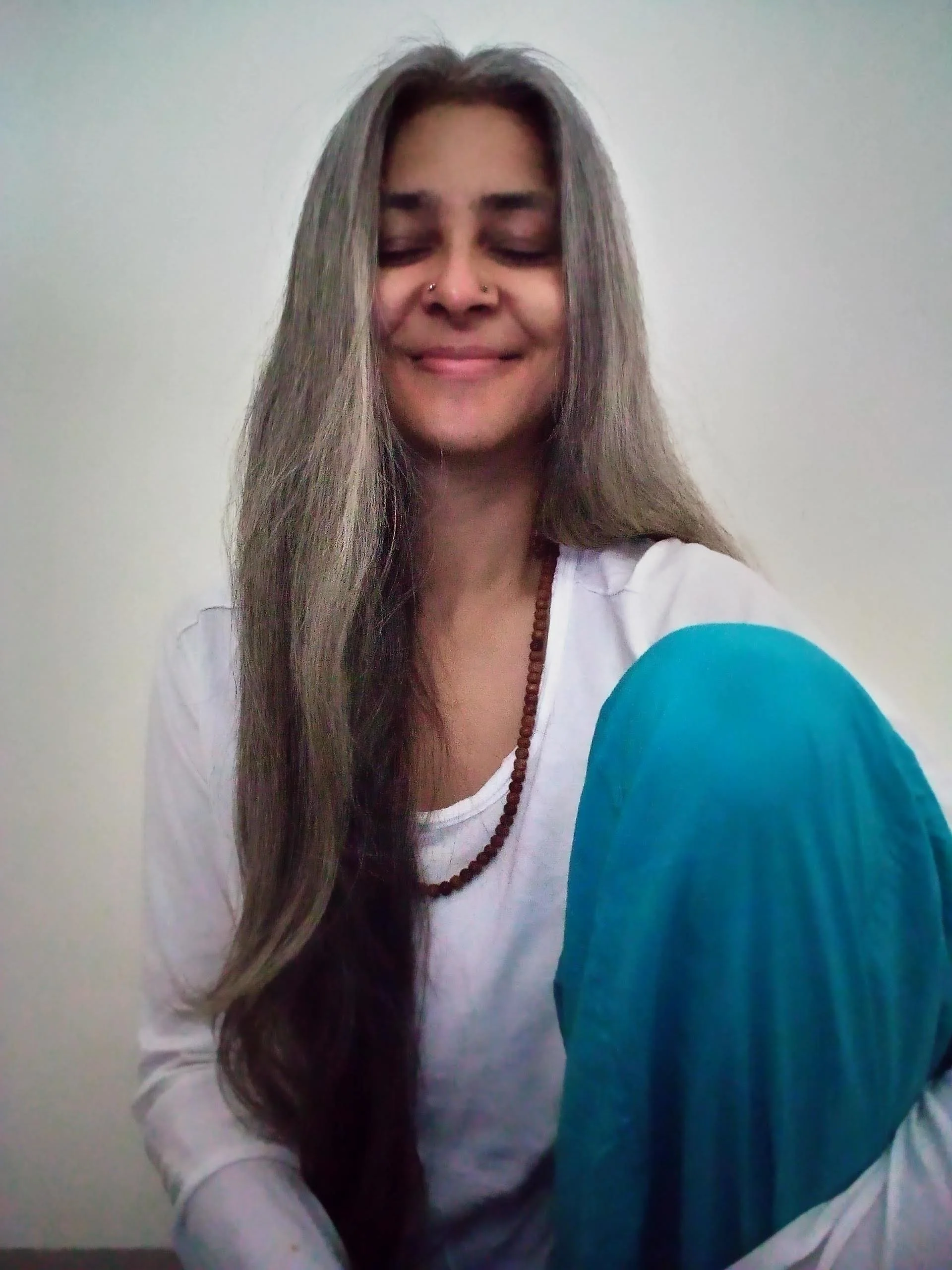Smiling person with long hair, wearing a white shirt and a turquoise garment, sitting with eyes closed, and a beaded necklace.