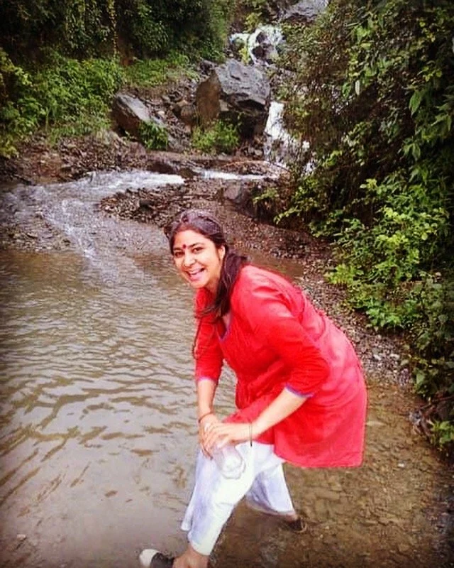 A person in a red top and white pants standing in shallow water near a small waterfall, surrounded by lush greenery.