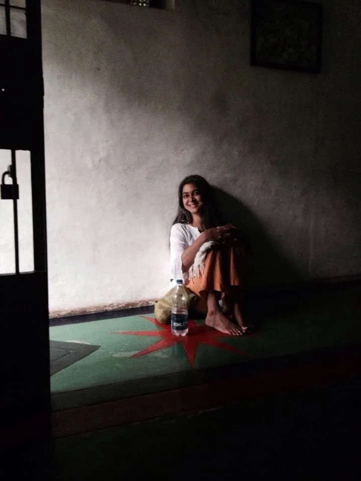 Woman sitting on floor, smiling, next to water bottle, in dimly lit room with simple decor.