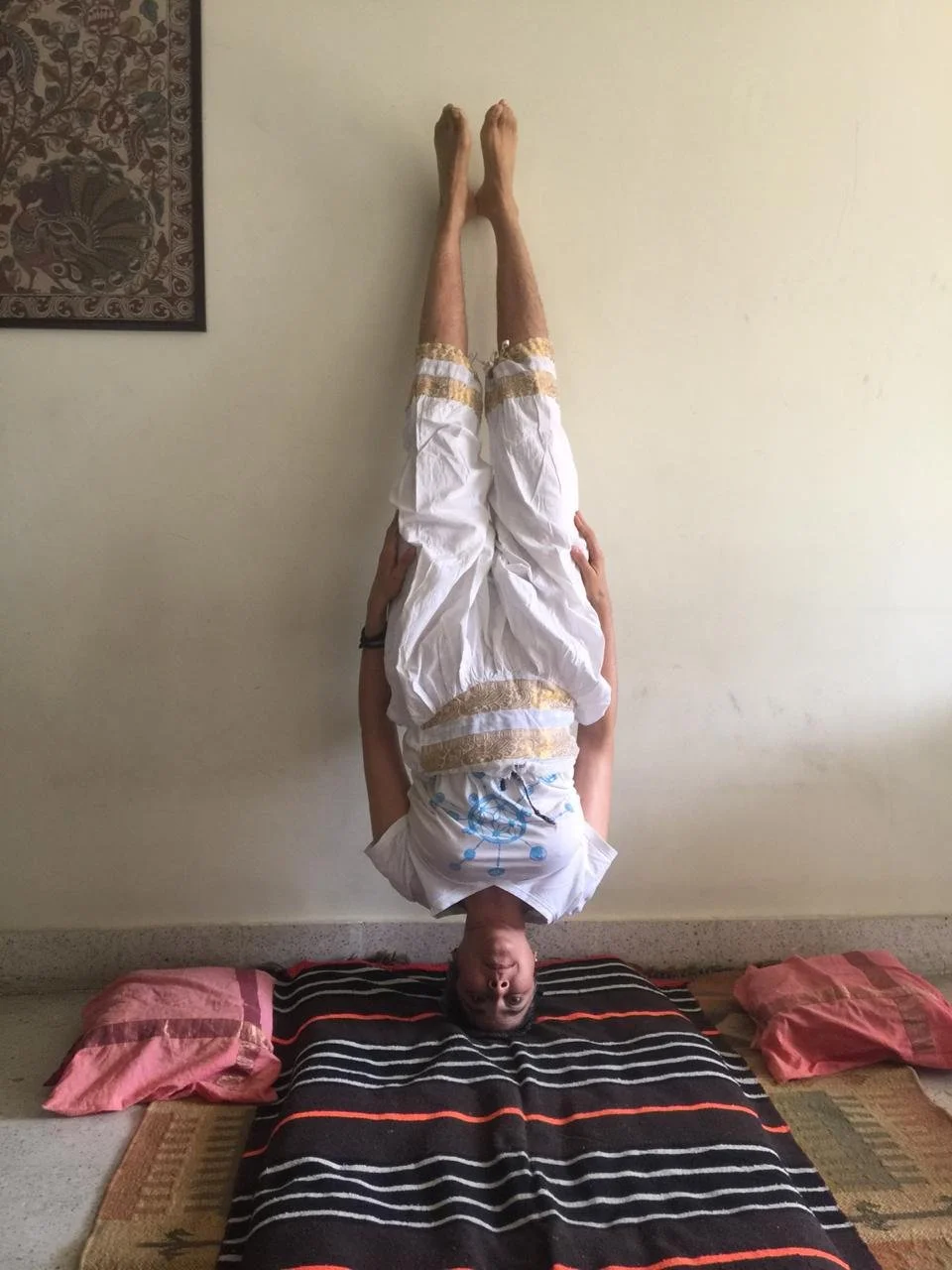 Person performing a headstand indoors, wearing white clothing, with a striped blanket on the floor and a painting on the wall.