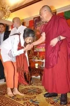 A woman bending to kiss the hand of a monk in a red robe, surrounded by others in a decorative room.