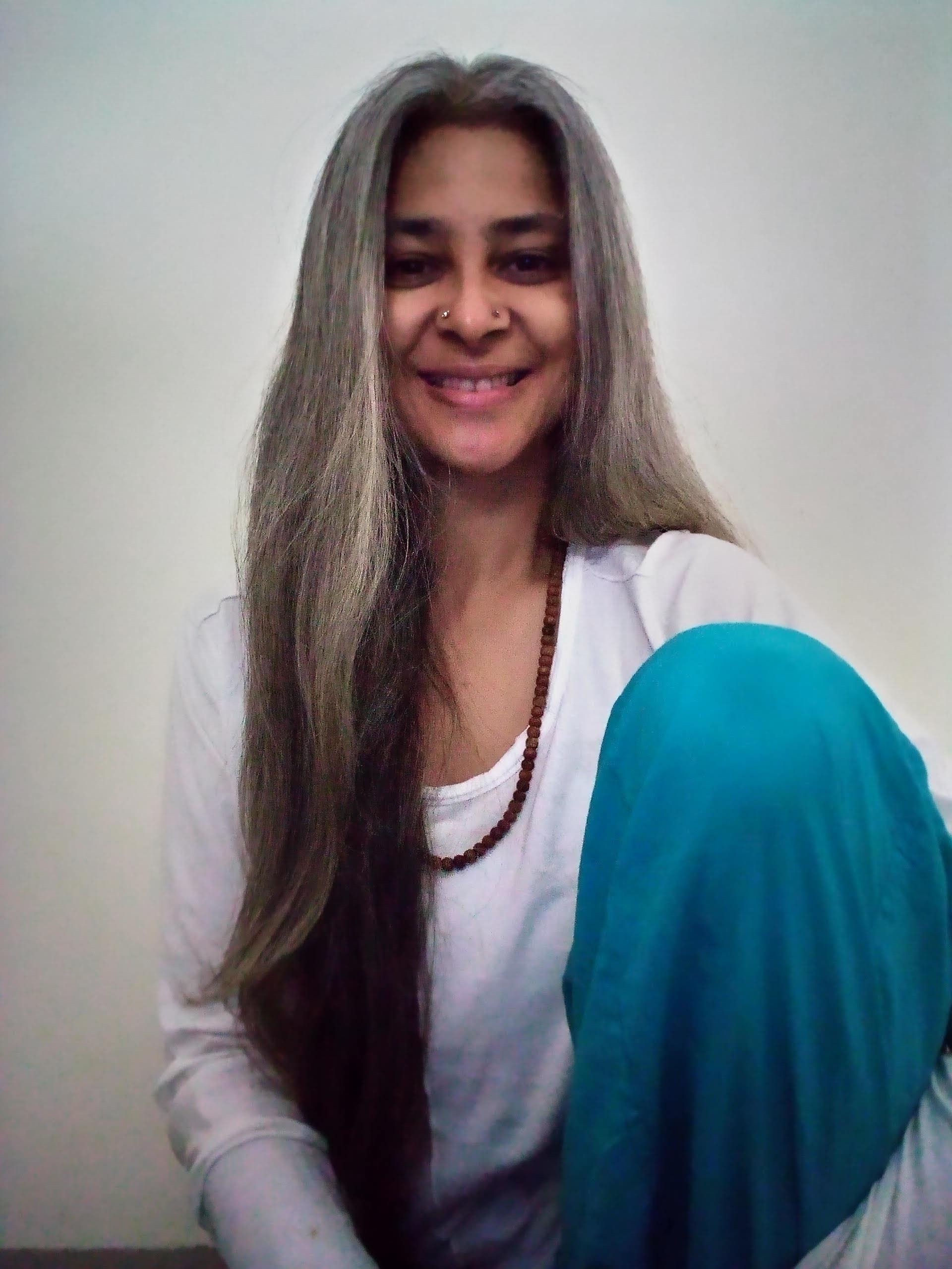 Smiling person with long gray hair, wearing a white long-sleeve shirt and turquoise pants, sitting indoors against a plain background.
