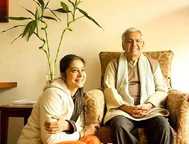 A smiling older man seated in an armchair next to a young woman sitting on the floor, against a background with green bamboo plants.