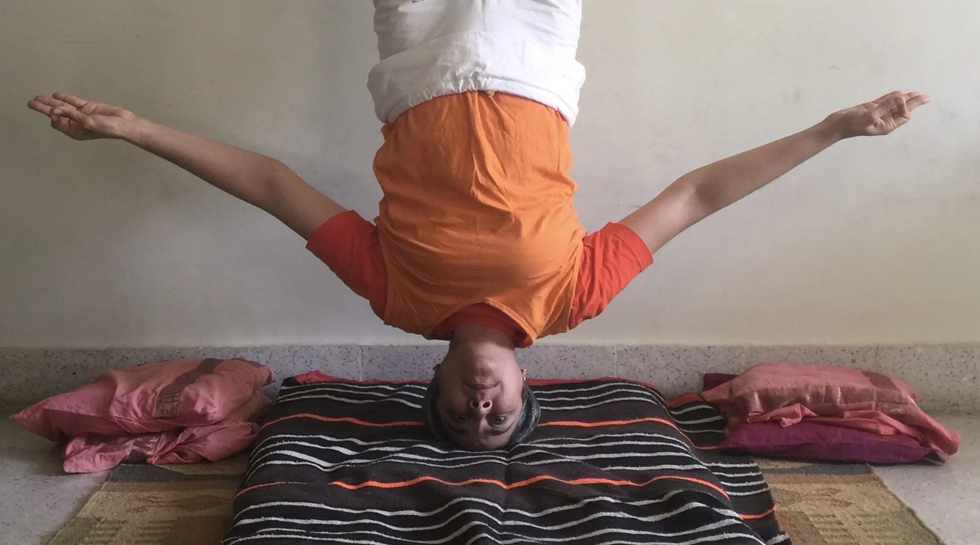 Person performing a headstand on a mat with arms spread, wearing an orange shirt and white pants.