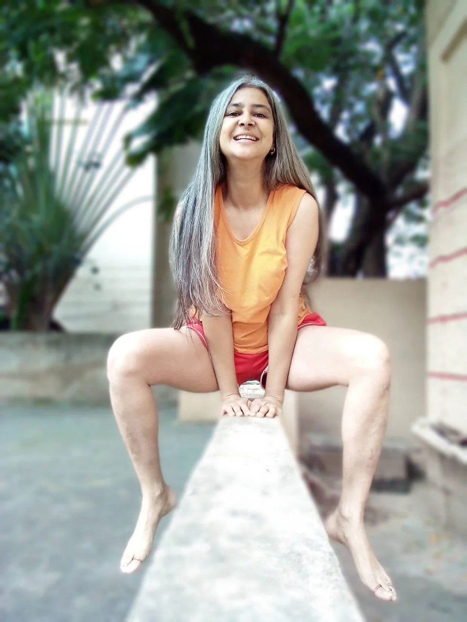 Person with long hair sitting outdoors on a concrete ledge, wearing an orange shirt and red shorts, smiling towards the camera, with trees in the background.