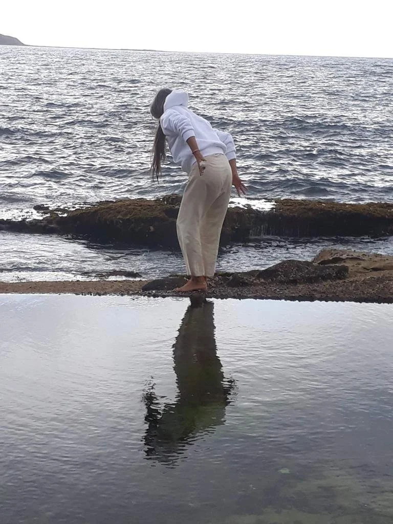 Person in white hoodie and beige pants bending over near a rocky shoreline, with their reflection in a water pool.