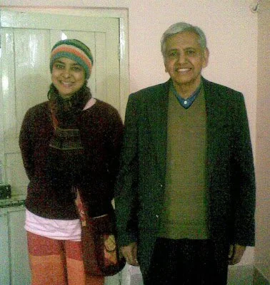 A man and woman smiling indoors, standing side by side. The woman is wearing a colorful knit hat, scarf, and layered clothing. The man is in a suit jacket and sweater.