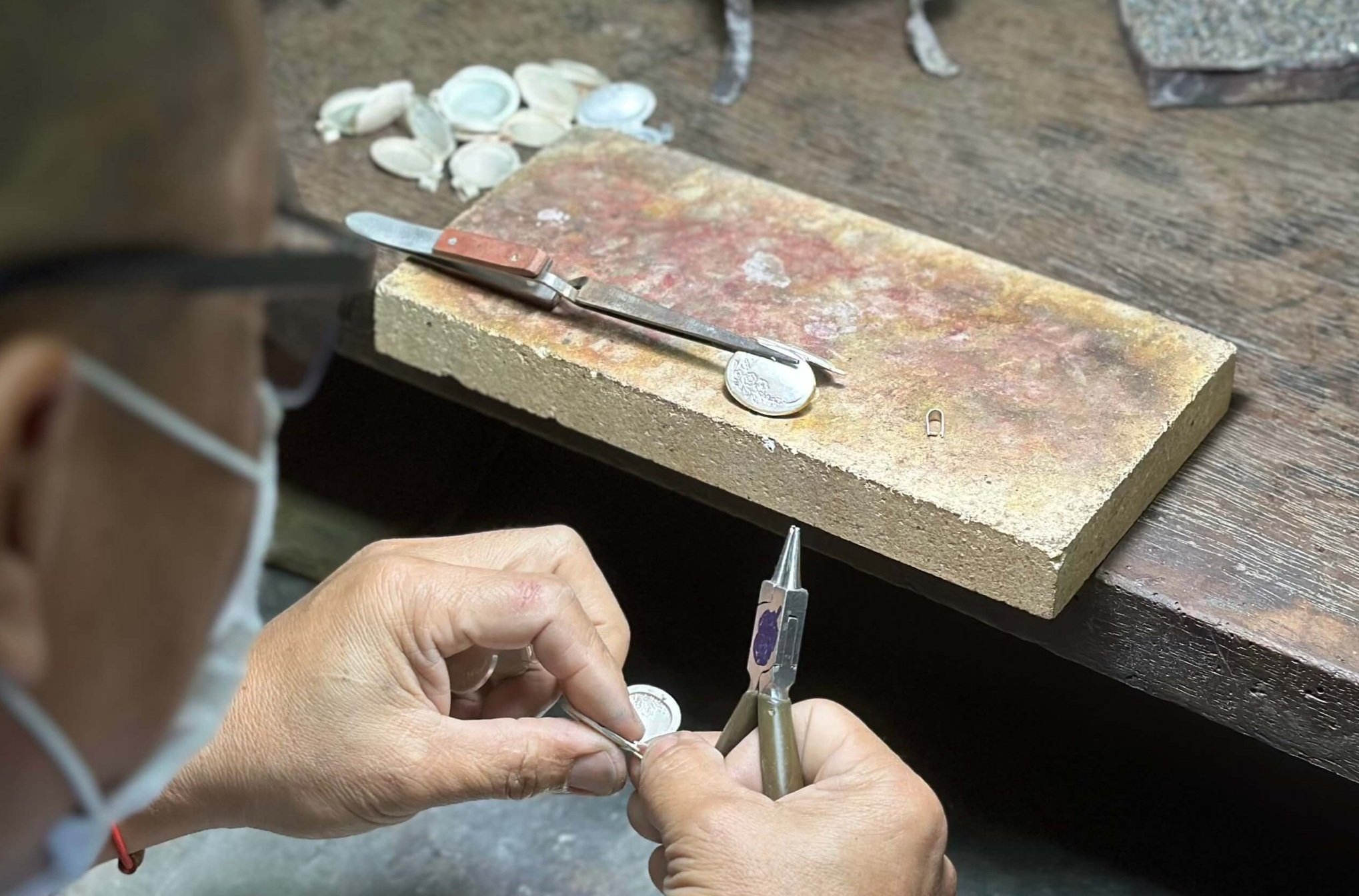 A person working on a coin with specialized tools in a workshop. The workspace has a brick surface and a wooden table with various metal pieces around.