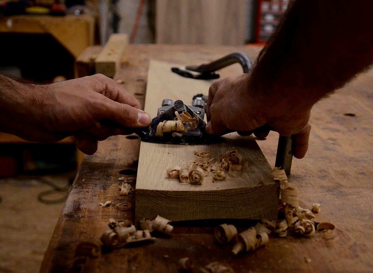 Two hands are planing a piece of wood with a hand planer, shavings are scattered on the workbench.