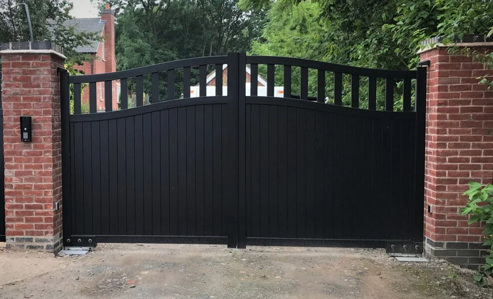 Black gated fence between two brick pillars with greenery in the background.