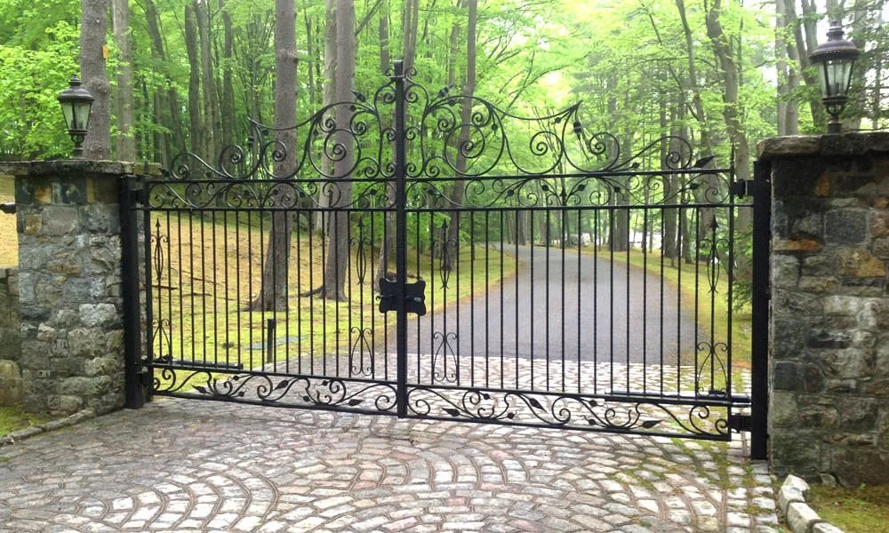 Black wrought iron gate with decorative scrolls, flanked by stone pillars with lanterns, opening to a paved driveway in a lush green forest.