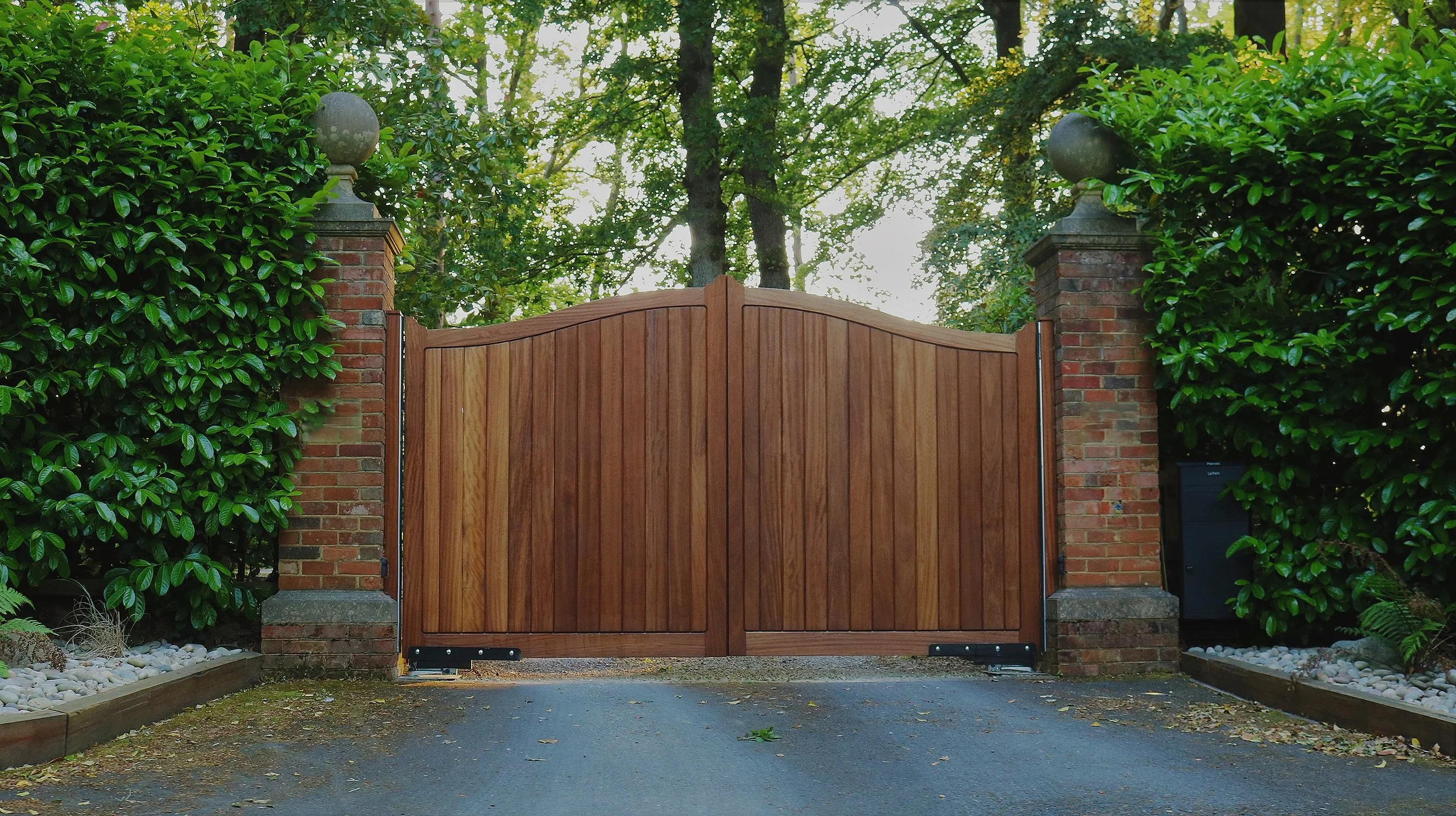 Wooden gate flanked by brick columns and leafy green bushes, with trees and sunlight in the background.
