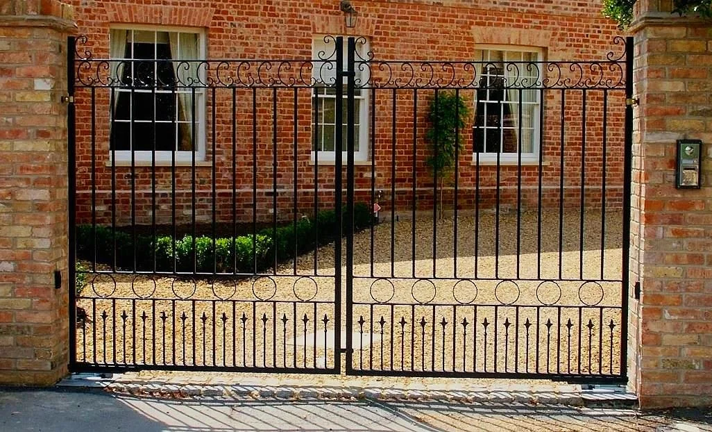 Black wrought iron gate with decorative scrollwork and circles, set between brick pillars in front of a brick house with two white-framed windows and a small landscaped garden.