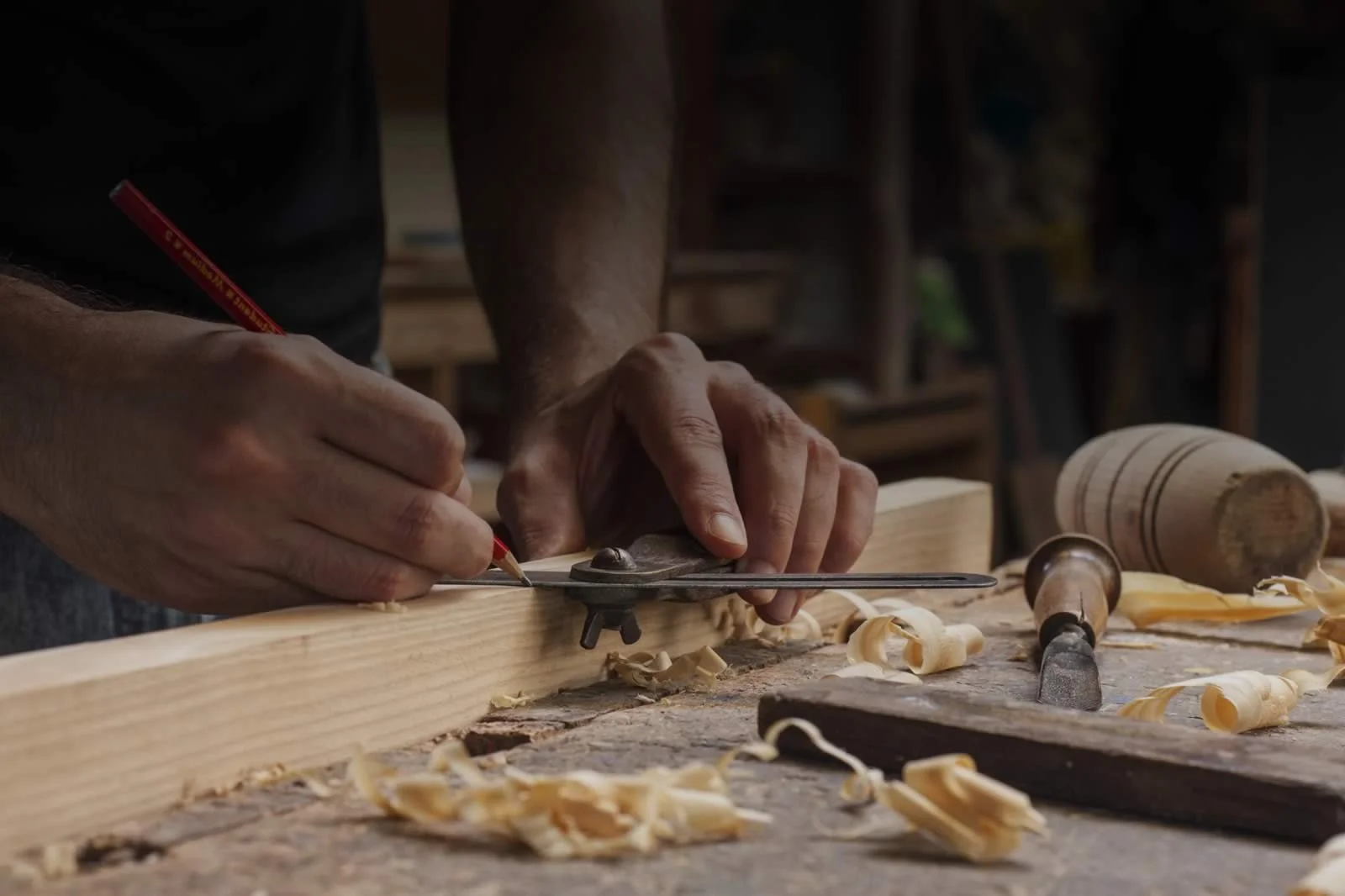 Close-up of a craftsman carving wood with a hand-held tool at a woodworking workshop, with wood shavings scattered on the work surface.