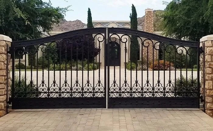 A black wrought iron gate with an arch at the top, flanked by stone pillars, leading to a driveway with a house and trees in the background.