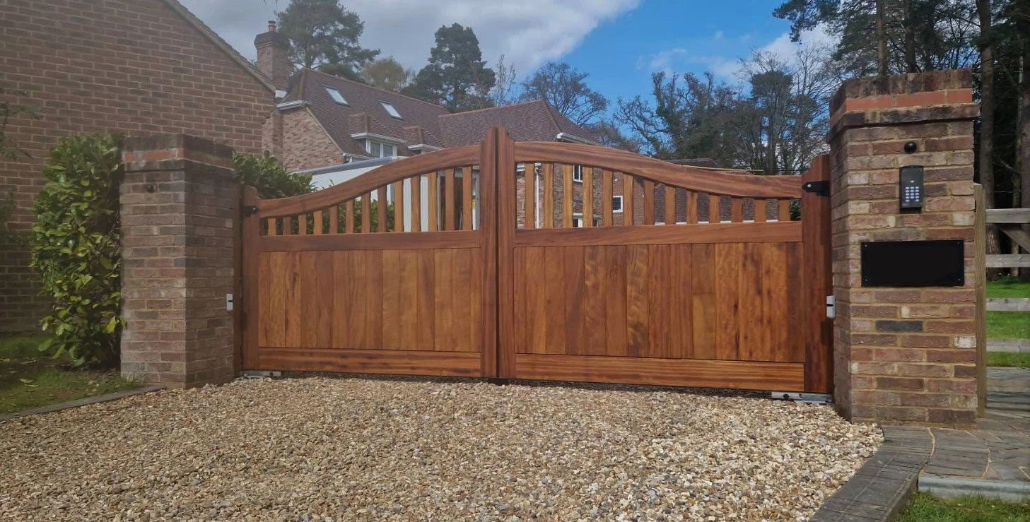 A wooden double gate with a decorative curved top, set between brick pillars on a gravel driveway, with a house and trees in the background.