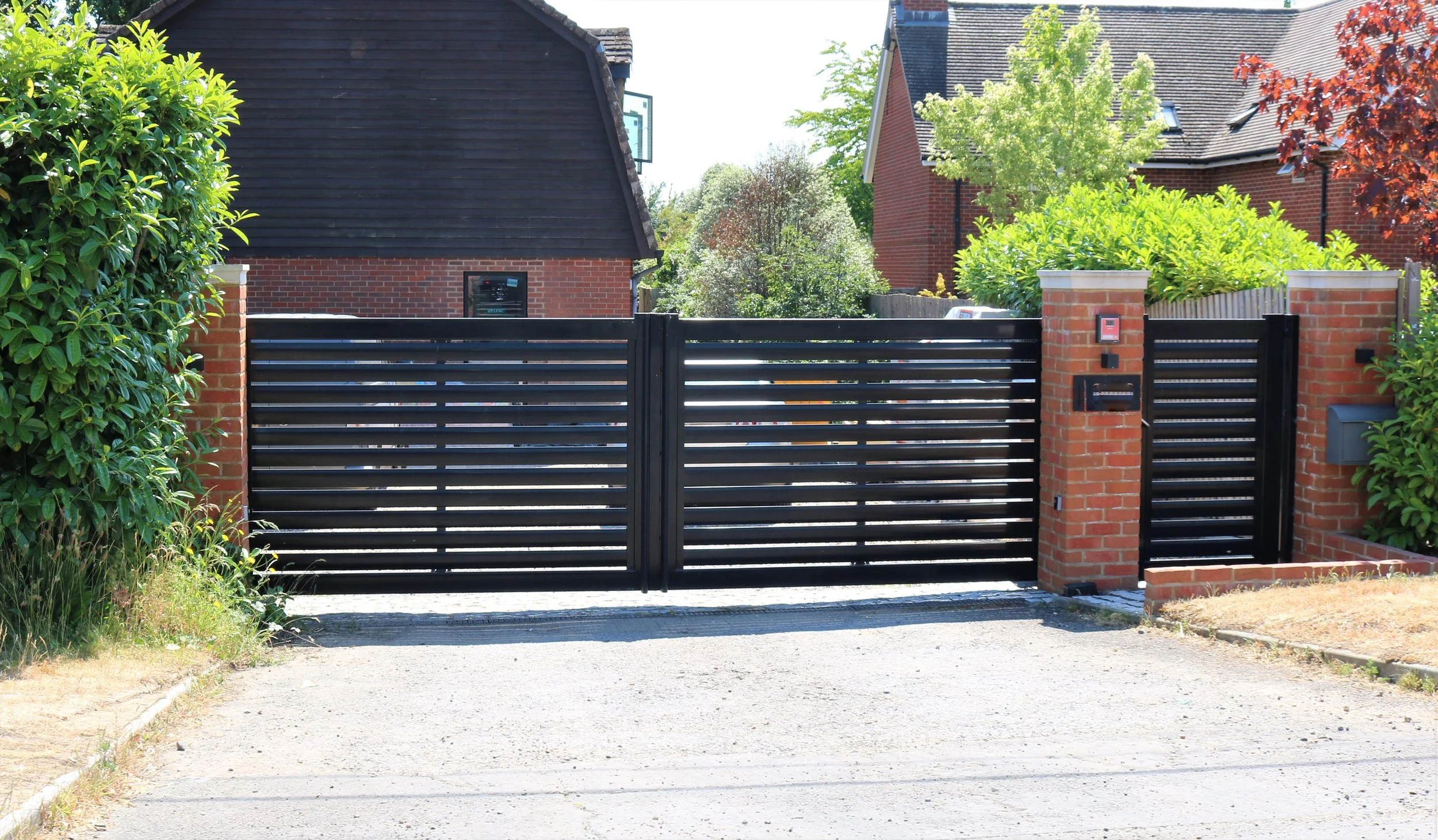 Black metal sliding gate with brick pillars on each side, leading to a residential driveway, with lush green trees and shrubs surrounding the brick houses.