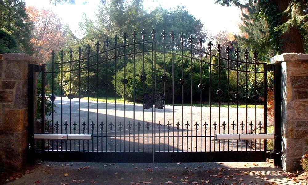 Black iron gate with stone pillars on each side, set in a driveway, surrounded by trees and greenery, with sunlight filtering through the foliage.