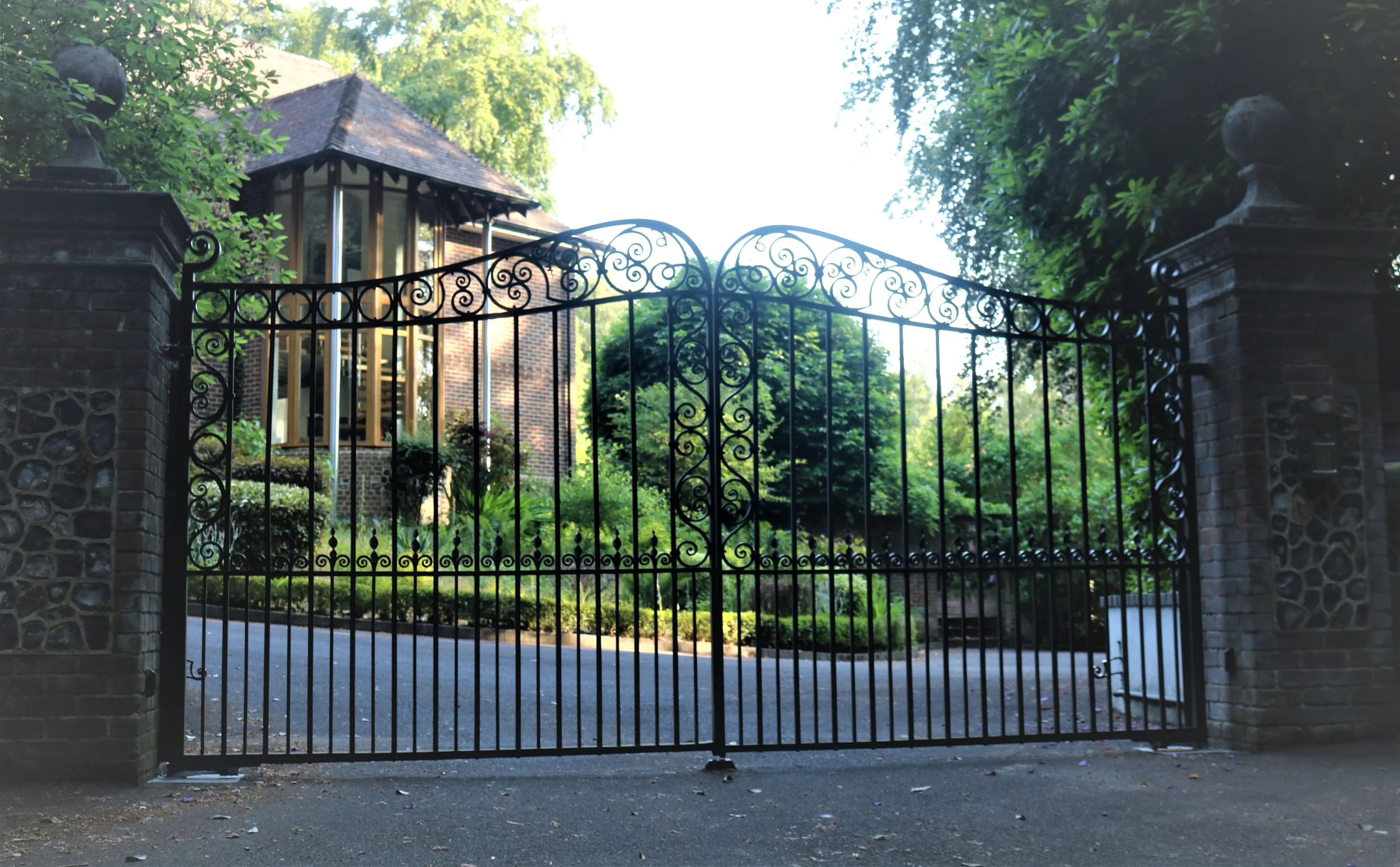 A black wrought iron gate with decorative swirls, flanked by brick and stone pillars, opens to a lush garden and a brick house with large windows, surrounded by green trees and plants.