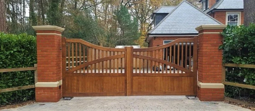 Wooden gate with brick pillars on each side, leading into a residential property with a brick house and trees.