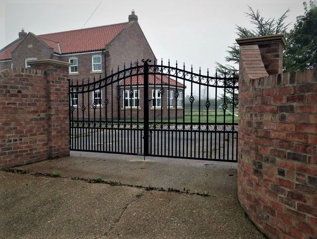 Black wrought iron gate between brick pillars, leading to a large brick house with a red-tiled roof surrounded by green lawn and trees.