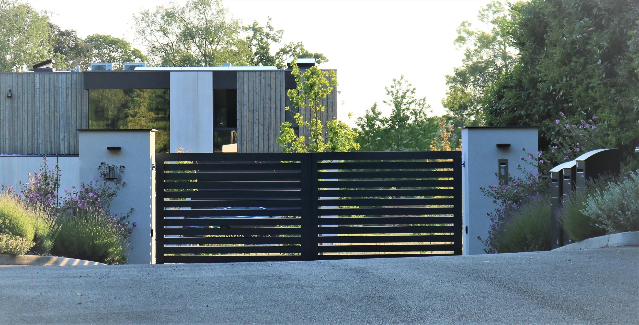 Modern house with wooden and white exterior, black horizontal slat gate, white walls with black accents, surrounded by flowering plants and green trees.