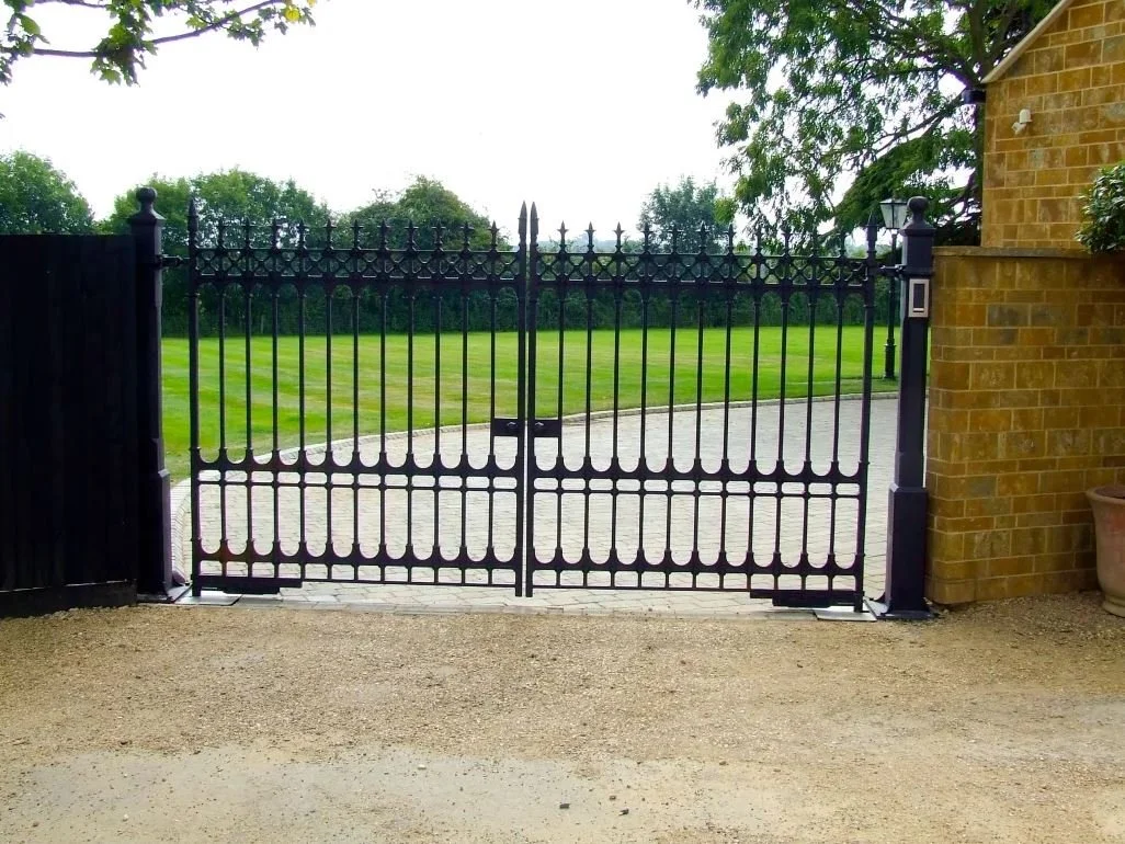 Black wrought iron gate leading to a green lawn and trees in the background, with a brick wall on the right and a black fence on the left.