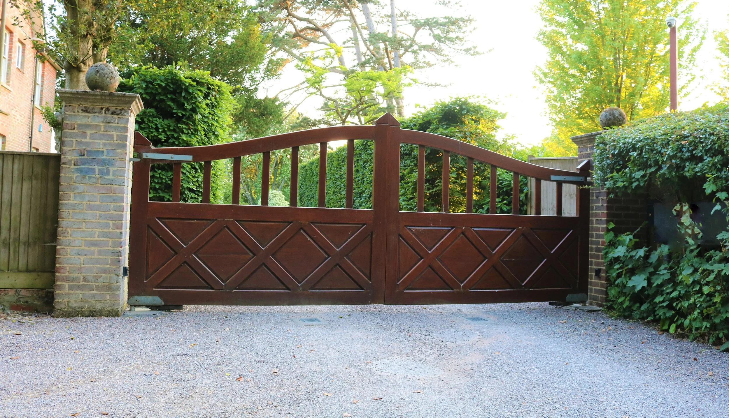 Residential driveway with a wooden gate, brick pillars with decorative spheres, surrounded by green bushes and trees.